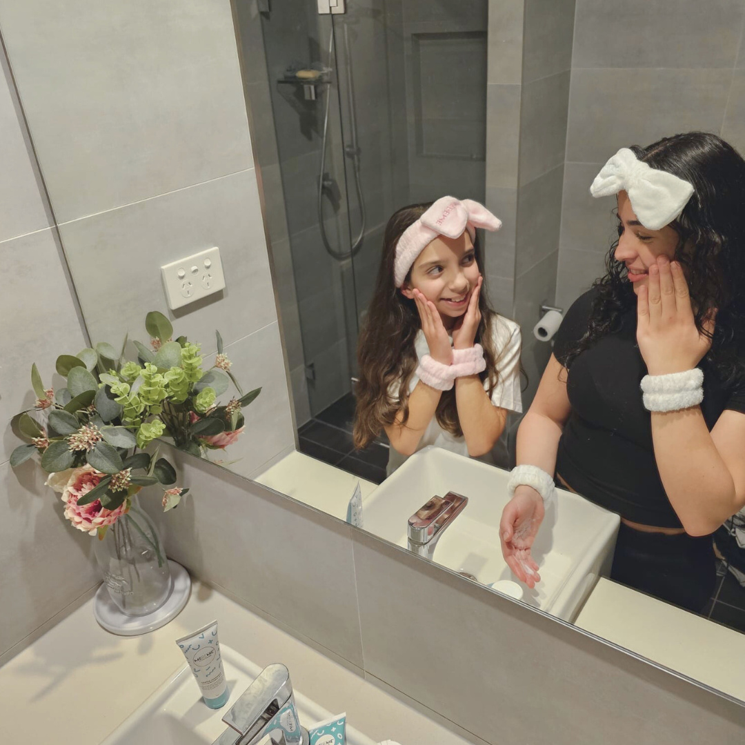 Two sisters applying teen moisturiser to their faces in a bathroom mirror.