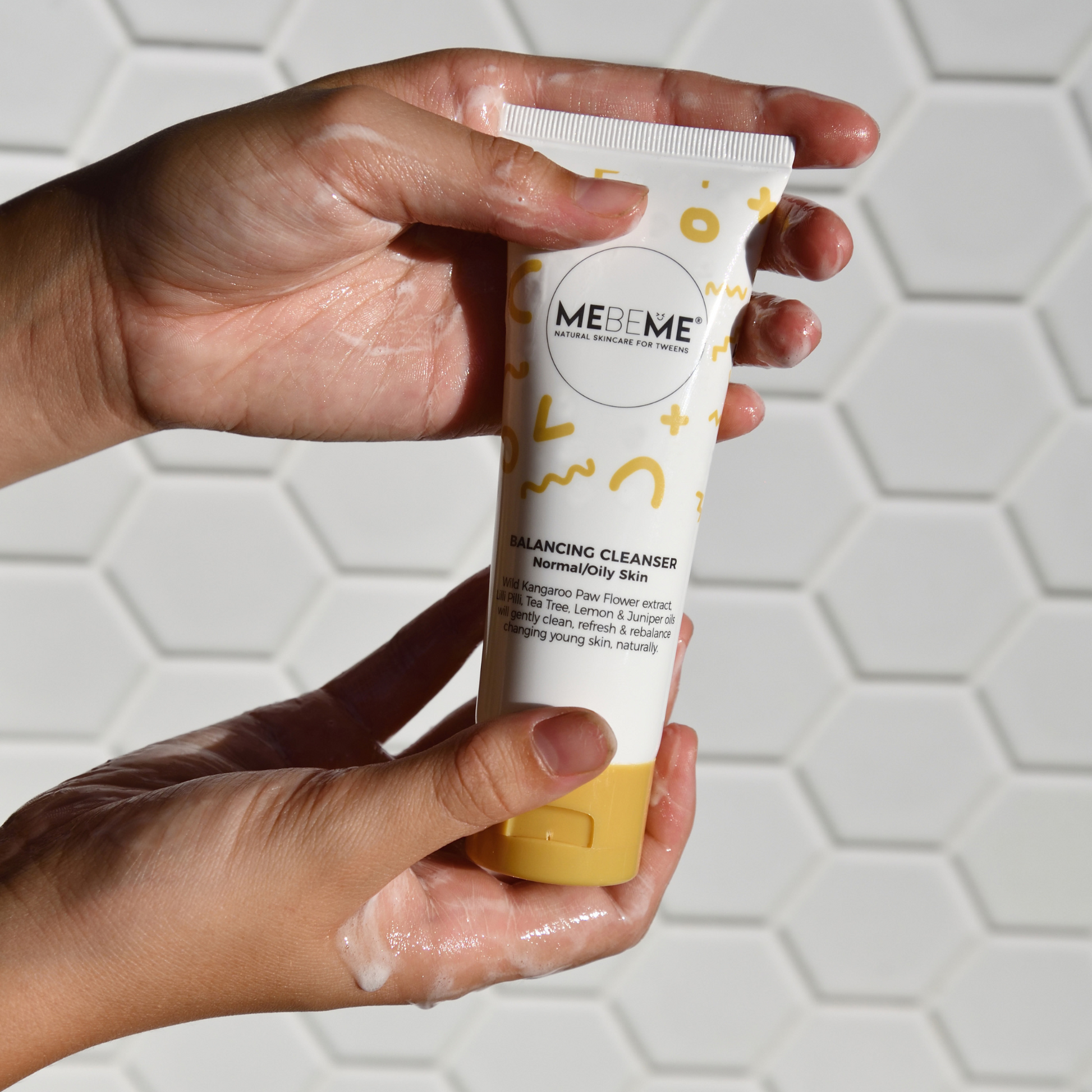 Hand holding a MEBEME skincare product with a white and yellow tube against a light geometric background