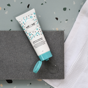 White tube of gentle cleanser for normal, sensitive or dry skin,  with blue and black squiggles. Tube has a blue lid which is open and the cleanser is pouring out onto a grey stone. Background is green with speckles and a white towel.