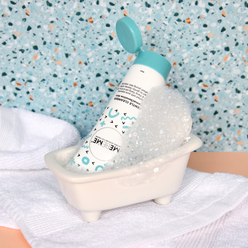 white tube of gentle cleanser for normal, sensitive or dry skin, with blue lid, with blue and black squiggles. Tube is sitting in a little bathtub with bubbles on a speckled background and white towel.
